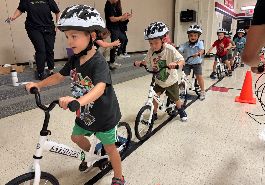  students ride balance bikes in one line in gym wearing helmets